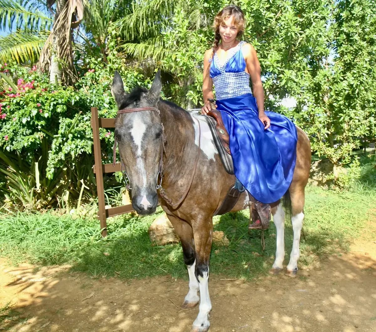 Riding a buckskin paint in a blue gown beside flowering bougainvillea